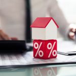 Representação de financiamento imobiliário: uma casa em miniatura colocada sobre um cubo com símbolo de porcentagem, simbolizando taxas de juros e crédito imobiliário, com profissionais ao fundo.
